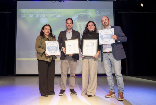 Four adults stand on a stage posing with framed certificates, signaling an awards ceremony; a screen behind them displays a banner about 100% renewable energy.
